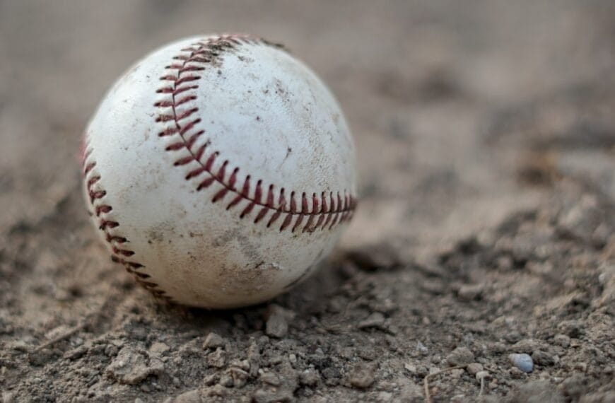 A worn baseball with visible dirt marks rests on a rough, sandy surface, reminiscent of the grit endured by baseball players. The red stitching stands prominent against the white of the ball. The image captures a close-up view, highlighting the texture of both the ball and the ground.
