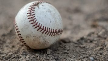 A worn baseball with visible dirt marks rests on a rough, sandy surface, reminiscent of the grit endured by baseball players. The red stitching stands prominent against the white of the ball. The image captures a close-up view, highlighting the texture of both the ball and the ground.