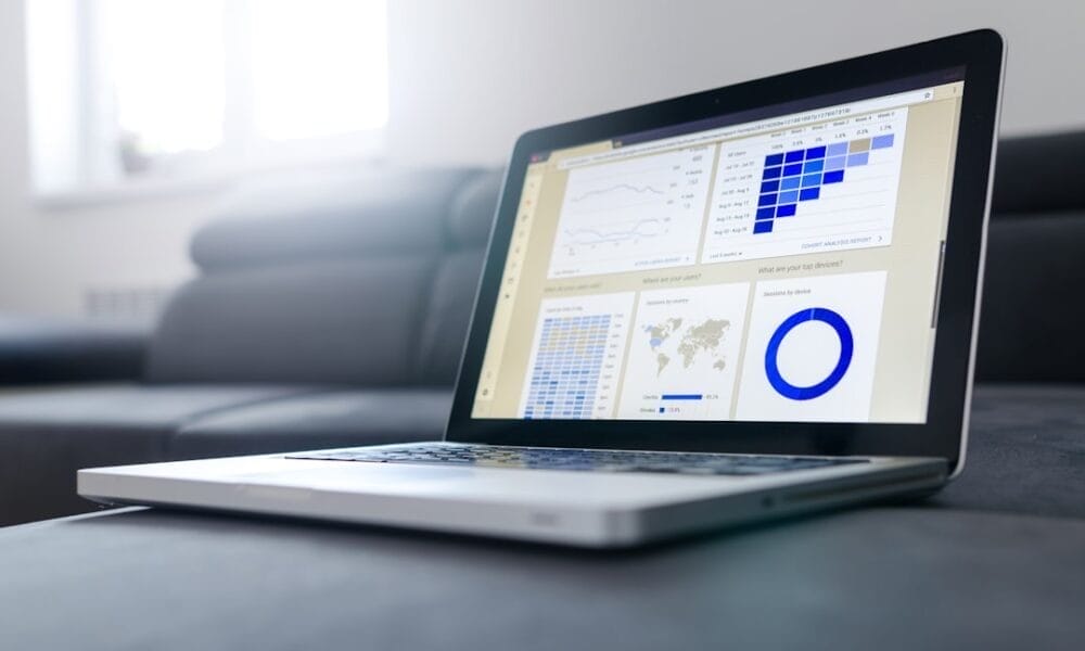 An open laptop rests on a couch, displaying statistical charts and graphs that delve into the intuition in sports analysis. The screen features a bar chart, line graph, world map, and circular graph. Sunlight streams through a nearby window, illuminating the scene with clarity.