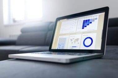 An open laptop rests on a couch, displaying statistical charts and graphs that delve into the intuition in sports analysis. The screen features a bar chart, line graph, world map, and circular graph. Sunlight streams through a nearby window, illuminating the scene with clarity.