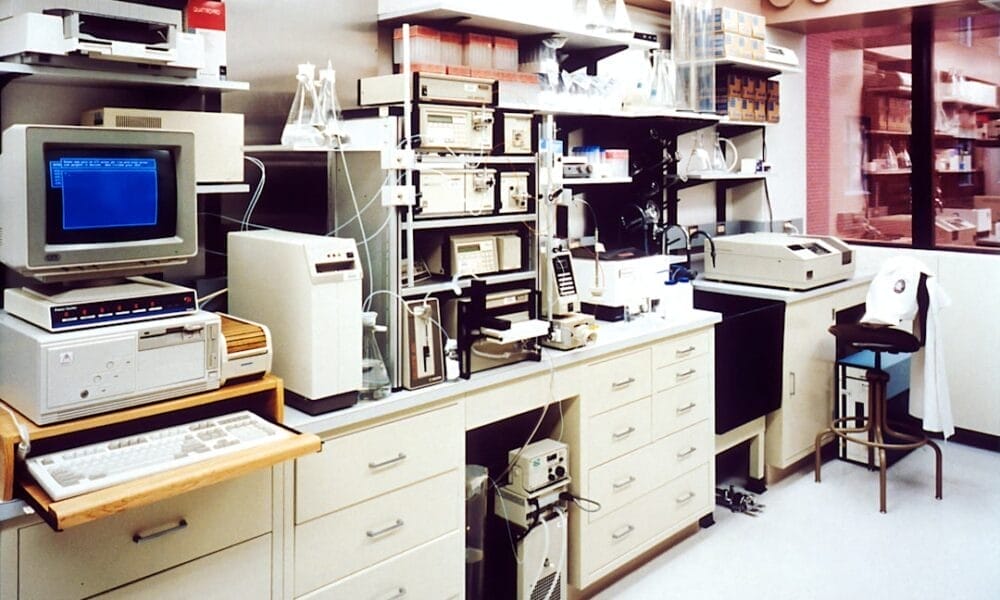 A retro laboratory features various scientific equipment and computers, including a CRT monitor displaying blue text. Shelves hold glassware and boxes, perhaps analyzing statistics in sports performance. The room has white cabinets and a fluorescent-lit, clean environment.