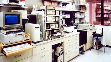 A retro laboratory features various scientific equipment and computers, including a CRT monitor displaying blue text. Shelves hold glassware and boxes, perhaps analyzing statistics in sports performance. The room has white cabinets and a fluorescent-lit, clean environment.