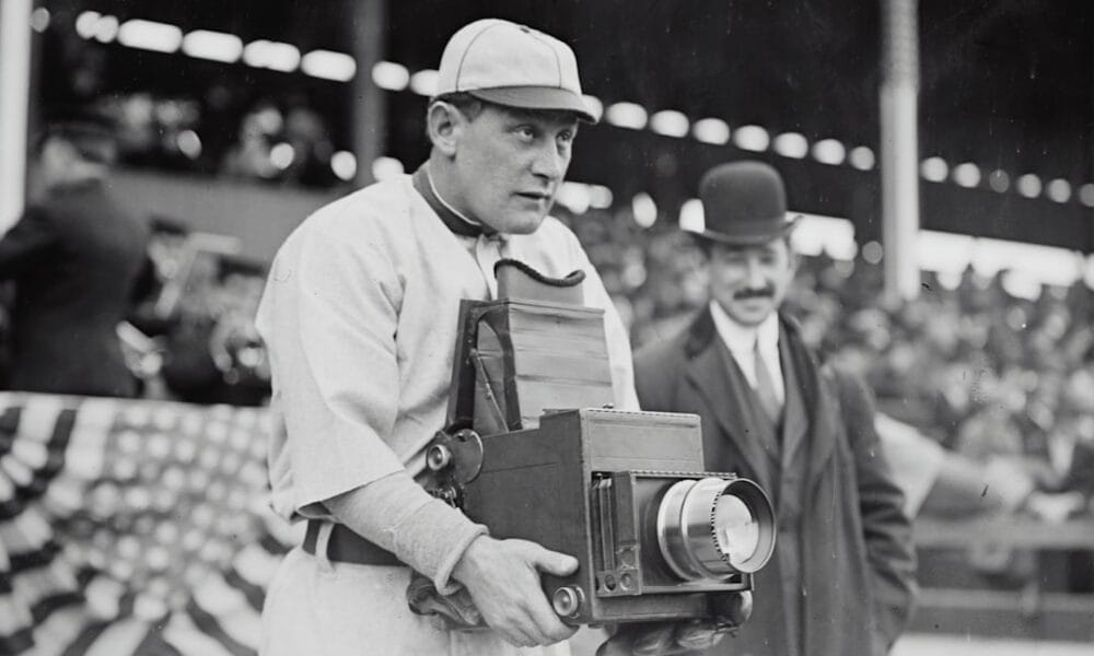 A man in an old-style baseball uniform, reminiscent of the early Baseball Eras, holds a large, vintage camera. Behind him, a man in a suit and bowler hat stands. In the background, blurred spectators and a draped American flag add to the nostalgic scene.