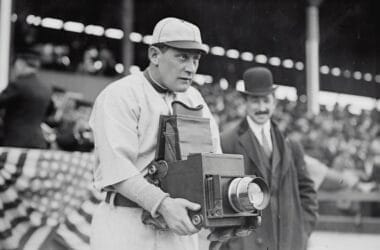 A man in an old-style baseball uniform, reminiscent of the early Baseball Eras, holds a large, vintage camera. Behind him, a man in a suit and bowler hat stands. In the background, blurred spectators and a draped American flag add to the nostalgic scene.