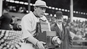 A man in an old-style baseball uniform, reminiscent of the early Baseball Eras, holds a large, vintage camera. Behind him, a man in a suit and bowler hat stands. In the background, blurred spectators and a draped American flag add to the nostalgic scene.