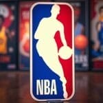 A large 3D NBA logo dominates the center of the image, featuring a basketball player silhouette in white against a vivid red and blue backdrop. Blurred colorful posters depicting iconic 90s Basketball on the NBA hang in the background, adding a nostalgic flair.