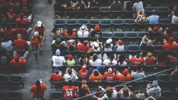 Aerial view of a crowd in stadium seating showcases fan loyalty as most people wear red shirts and hats. A mix of other colors is visible, with some seats empty. The setting appears to be a sports event, with attendees mostly seated and a few standing or walking.