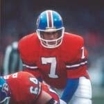 A football player in a bright orange Denver Broncos uniform and blue helmet, number 7, crouches behind a teammate. Focused and ready to play, John Elway sports a white chinstrap and blue sleeves under his jersey while the background is blurred.
