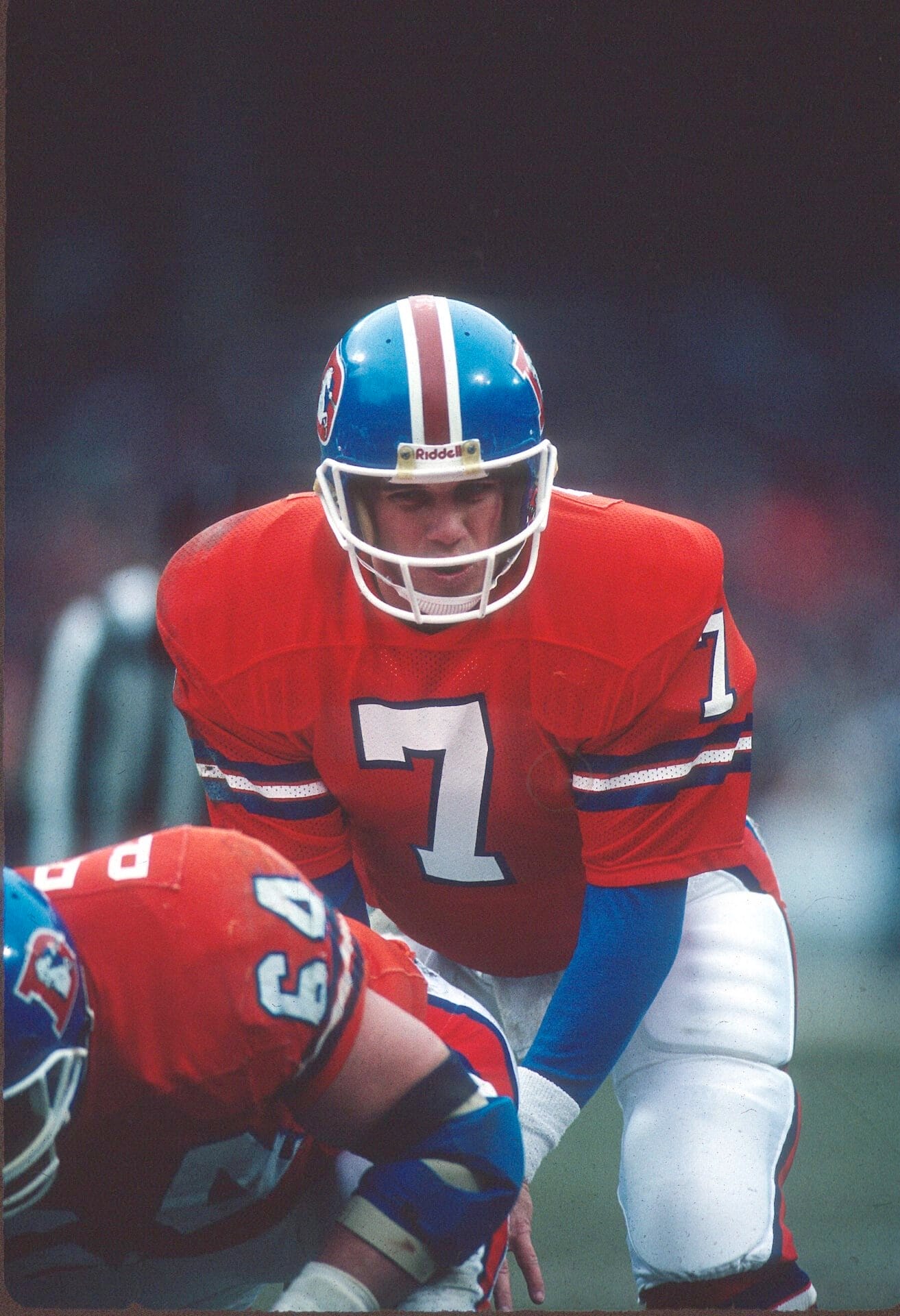 A football player in a bright orange Denver Broncos uniform and blue helmet, number 7, crouches behind a teammate. Focused and ready to play, John Elway sports a white chinstrap and blue sleeves under his jersey while the background is blurred.