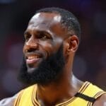 A basketball player in a yellow jersey smiles during a game. He has a full beard, short hair, and visible sweat on his forehead. The blurred stadium lights hint at the intensity and Hot Takes in Sports that surround every play.