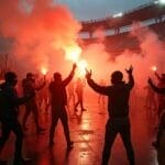 A group of people, some wearing hoodies and masks, stand in a stadium holding up red flares that emit thick smoke, creating a dramatic, glowing atmosphere. The wet ground reflects the light, and stadium seats are visible in the background.