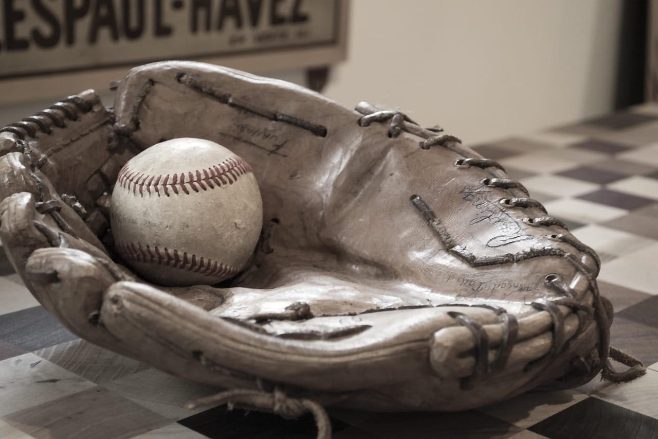 A worn brown leather baseball glove with visible signatures cradles a scuffed baseball from the Live Ball Era on a checkered wooden table. In the blurred background, part of a vintage sign with faded lettering is visible on the wall.