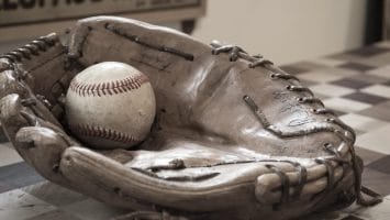 A worn brown leather baseball glove with visible signatures cradles a scuffed baseball from the Live Ball Era on a checkered wooden table. In the blurred background, part of a vintage sign with faded lettering is visible on the wall.