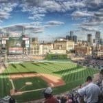 A panoramic view of a baseball stadium filled with fans, the green field and scoreboard evoke memories of the steroid era. The city skyline rises behind the stands under a partly cloudy sky as sunlight casts gentle shadows across the scene.