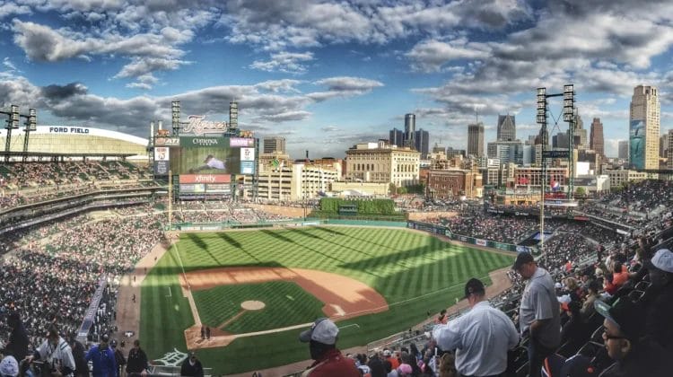 A panoramic view of a baseball stadium filled with fans, the green field and scoreboard evoke memories of the steroid era. The city skyline rises behind the stands under a partly cloudy sky as sunlight casts gentle shadows across the scene.