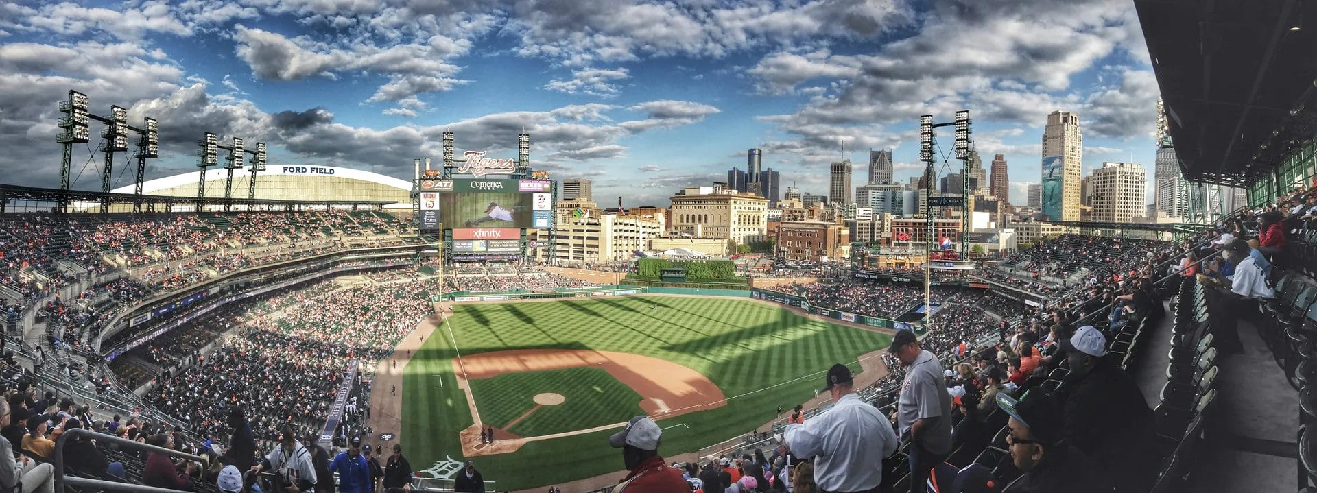 A panoramic view of a baseball stadium filled with fans, the green field and scoreboard evoke memories of the steroid era. The city skyline rises behind the stands under a partly cloudy sky as sunlight casts gentle shadows across the scene.