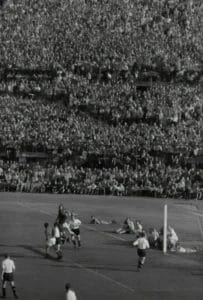 Black-and-white photo of a packed stadium during a soccer match. Players in light and dark jerseys crowd by the goal as the goalkeeper jumps to catch the ball. Thousands of spectators fill the stands, watching the action closely.