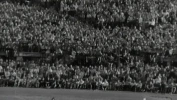 Black-and-white photo of a packed stadium during a soccer match. Players in light and dark jerseys crowd by the goal as the goalkeeper jumps to catch the ball. Thousands of spectators fill the stands, watching the action closely.