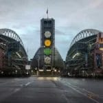 A wide-angle view of Lumen Field stadium in Seattle, home to some of the NFL’s fiercest football rivalries, shows its twin curved roofs and central tower with clocks. The empty stadium has wet pavement in front and a cloudy sky overhead.