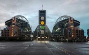 A wide-angle view of Lumen Field stadium in Seattle, home to some of the NFL’s fiercest football rivalries, shows its twin curved roofs and central tower with clocks. The empty stadium has wet pavement in front and a cloudy sky overhead.