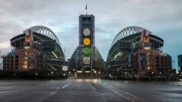 A wide-angle view of Lumen Field stadium in Seattle, home to some of the NFL’s fiercest football rivalries, shows its twin curved roofs and central tower with clocks. The empty stadium has wet pavement in front and a cloudy sky overhead.