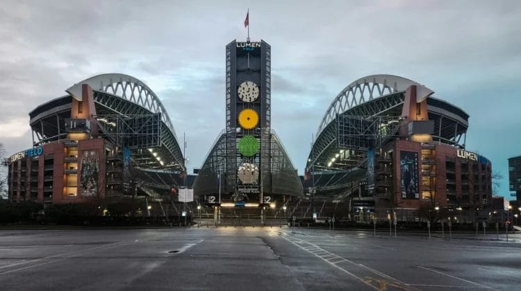 A wide-angle view of Lumen Field stadium in Seattle, home to some of the NFL’s fiercest football rivalries, shows its twin curved roofs and central tower with clocks. The empty stadium has wet pavement in front and a cloudy sky overhead.