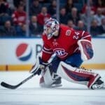 A hockey goalie in a red Montreal Canadiens jersey and full gear crouches on the ice, gripping a black stick and watching play—an ideal scene for hockey analytics, with blurred spectators in red and rinkside advertisements completing the backdrop.