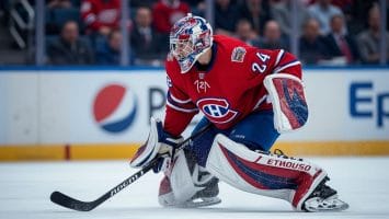 A hockey goalie in a red Montreal Canadiens jersey and full gear crouches on the ice, gripping a black stick and watching play—an ideal scene for hockey analytics, with blurred spectators in red and rinkside advertisements completing the backdrop.