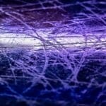Close-up of a purple-lit ice rink surface, covered with intersecting skate marks and scratches. The textured effect hints at the history of NHL games played, highlighting the roughness and chaotic patterns etched into the ice. No people or objects are visible.
