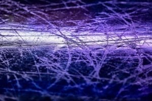 Close-up of a purple-lit ice rink surface, covered with intersecting skate marks and scratches. The textured effect hints at the history of NHL games played, highlighting the roughness and chaotic patterns etched into the ice. No people or objects are visible.