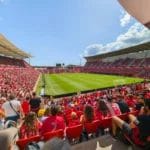 A packed football stadium with thousands of fans in red fills the stands. As a La Liga Expert would note, spectators face a green soccer field under sunny skies, with seating areas curving around the pitch and players visible on the field.