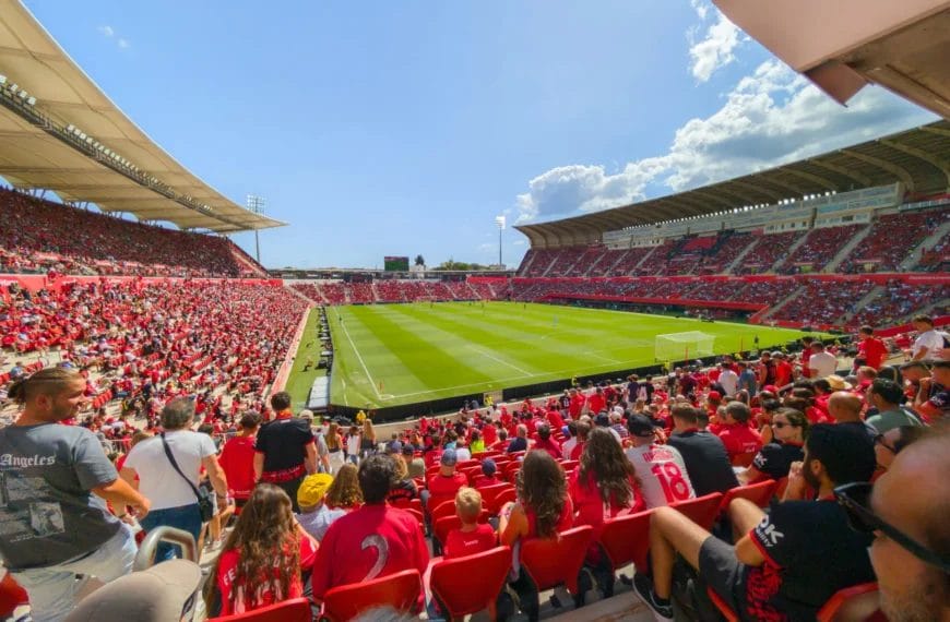 A packed football stadium with thousands of fans in red fills the stands. As a La Liga Expert would note, spectators face a green soccer field under sunny skies, with seating areas curving around the pitch and players visible on the field.