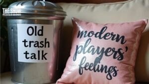A metal trash can labeled “Old Trash Talk” sits next to a pink glittery pillow with the words “modern players’ feelings” written in cursive, both resting on a beige couch.