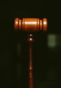 A close-up of a wooden judge’s gavel against a dark, blurred background. The gavel is centered and upright, highlighting its smooth, polished texture and symbolic association with law and justice.
