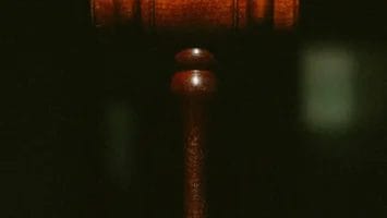 A close-up of a wooden judge’s gavel against a dark, blurred background. The gavel is centered and upright, highlighting its smooth, polished texture and symbolic association with law and justice.