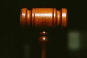 A close-up of a wooden judge’s gavel against a dark, blurred background. The gavel is centered and upright, highlighting its smooth, polished texture and symbolic association with law and justice.
