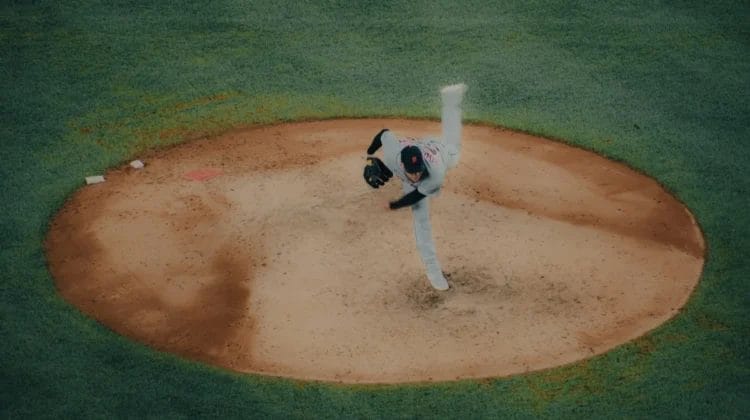 A baseball pitcher in a white uniform is captured mid-pitch atop a brown dirt mound, surrounded by green grass—a striking display of athleticism in the modern baseball era. His right leg is high and his arm extended, highlighting the game's dynamic motion.