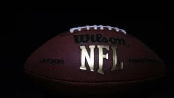 A close-up of an American football with "Wilson" and "NFL" in large gold letters on its brown, textured surface evokes the excitement of NFL contracts. The ball rests against a dark background, with white laces and stitching partially visible at the top.